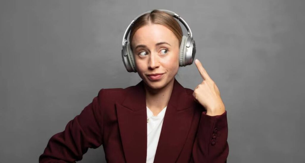 A woman wearing large headphones points upward, dressed in a burgundy blazer against a gray background.