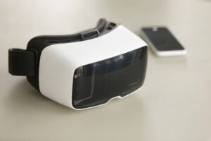 A white and black virtual reality headset rests on a light surface next to a black smartphone.