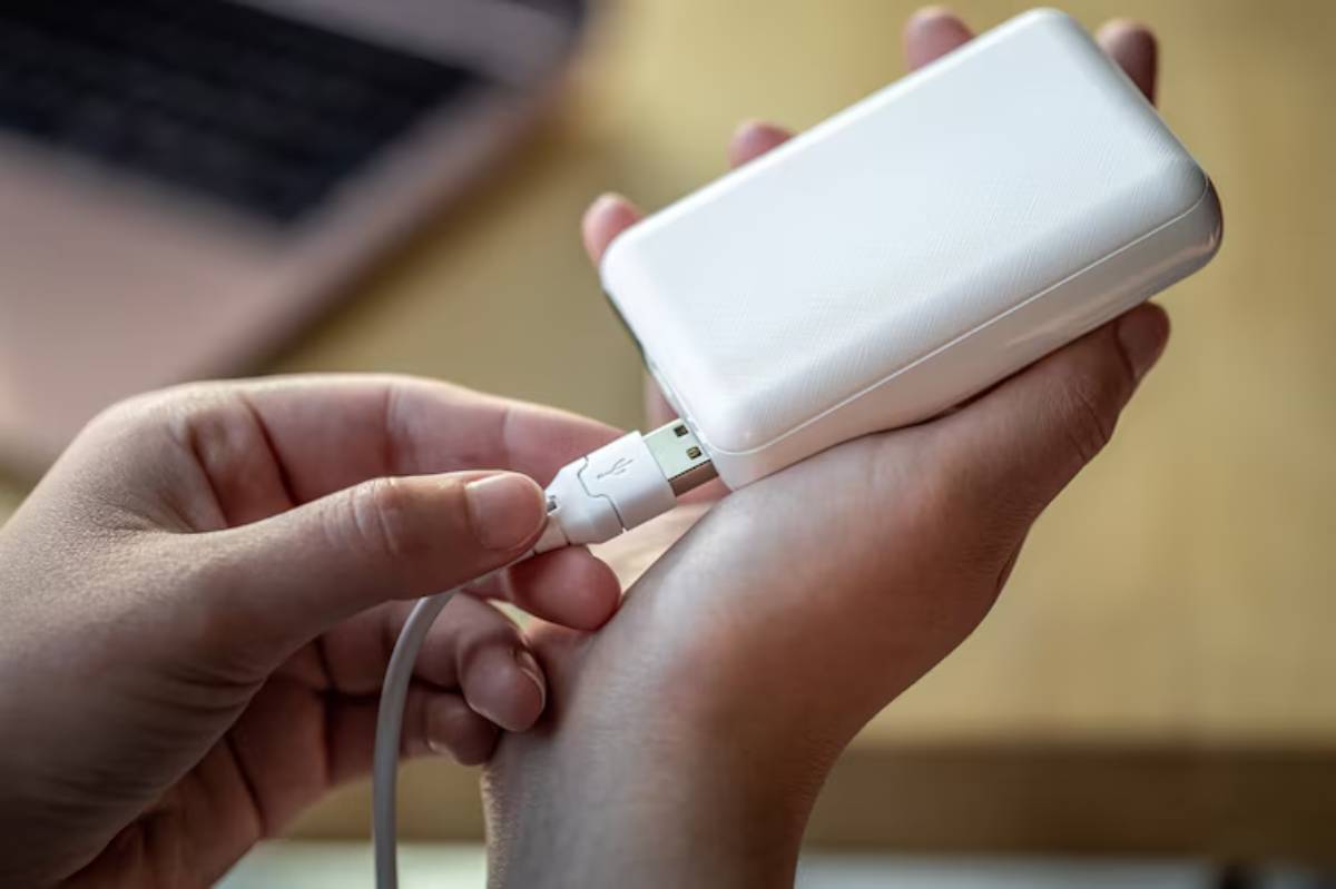 A person connects a white power bank to a charging cable, with a laptop in the background on a wooden table.