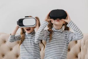 Two girls with long, curly hair wearing striped turtlenecks are enjoying virtual reality headsets while sitting on a plush couch.