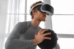 A person in a gray long-sleeve shirt holds a textured black ball while wearing a virtual reality headset in a bright room.