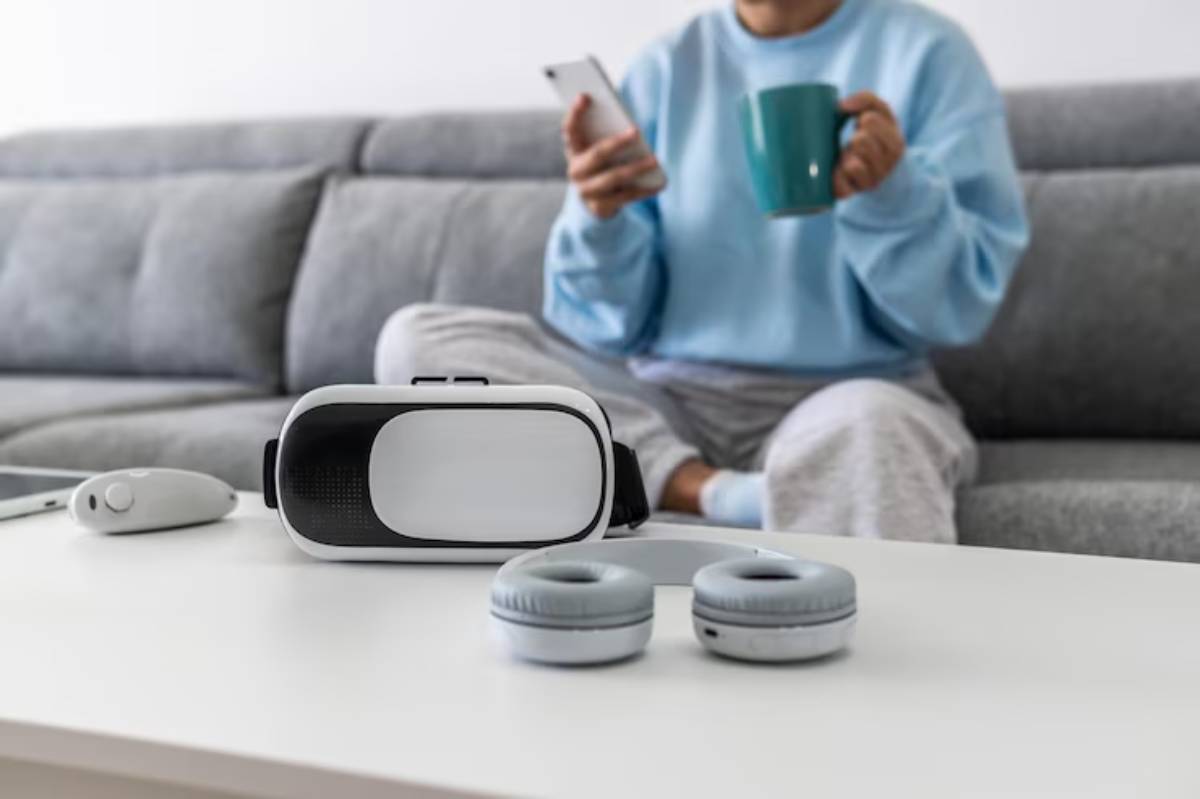 A person in a blue sweatshirt sits on a couch, holding a phone and a mug, with a VR headset and headphones on the table.