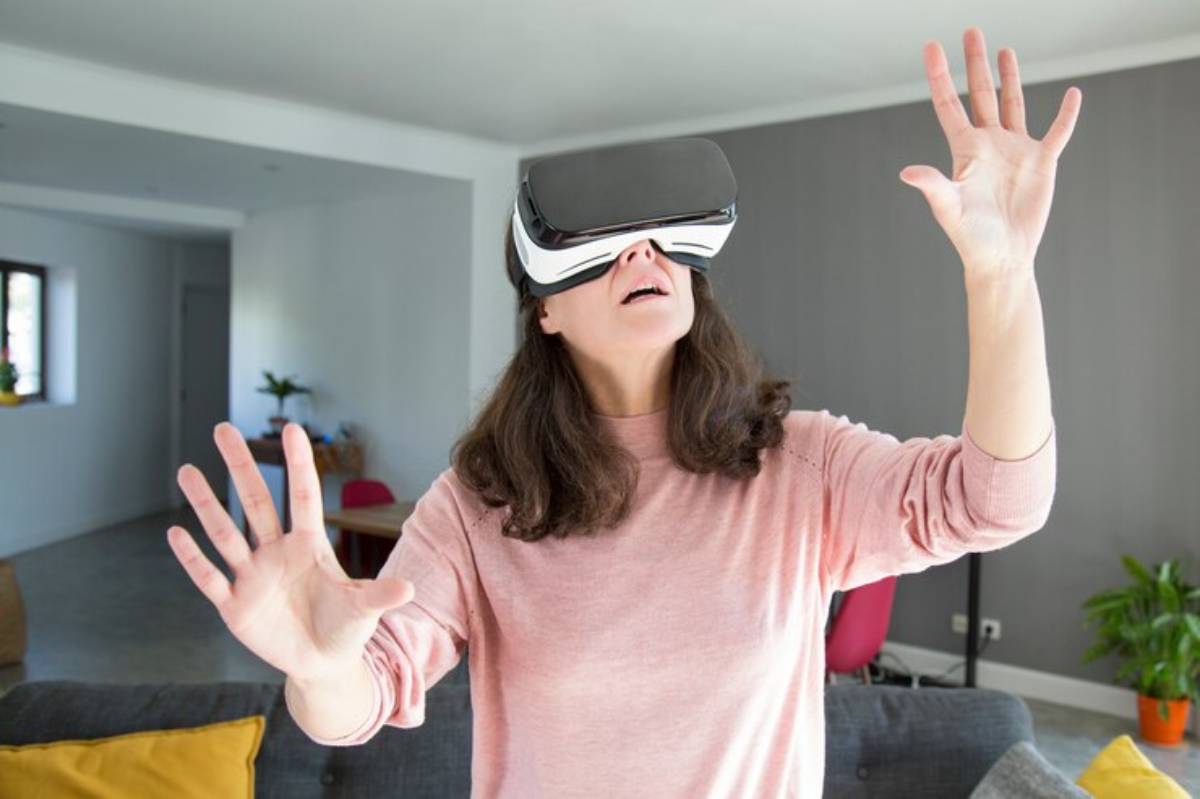 A person wearing virtual reality goggles stands in a modern living room, reaching out with both hands as if interacting with a virtual environment.