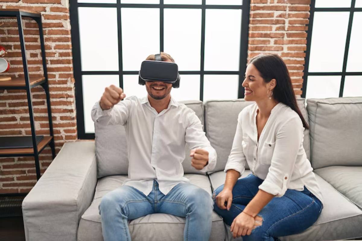A person wearing a VR headset sits on a couch, engaging enthusiastically, while another person observes nearby in a cozy setting.
