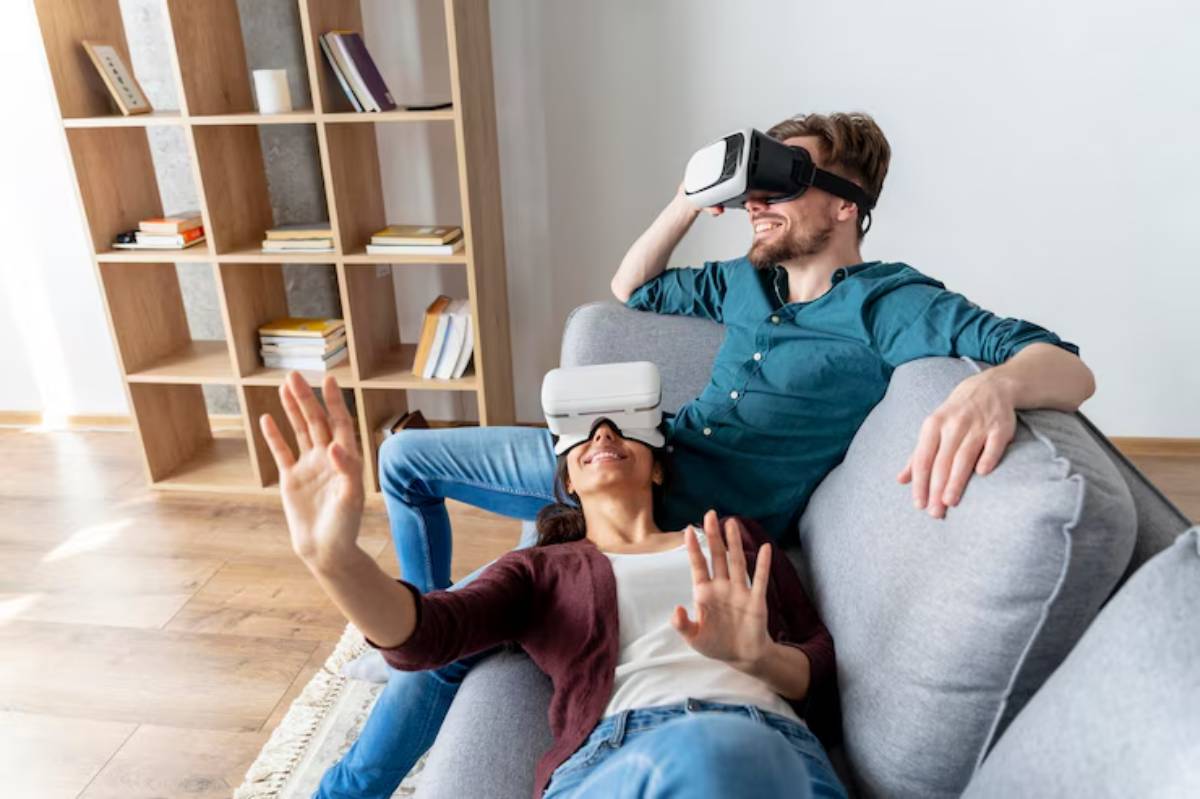 A couple relaxes on a couch, both wearing virtual reality headsets in a cozy living room with a wooden bookshelf in the background.