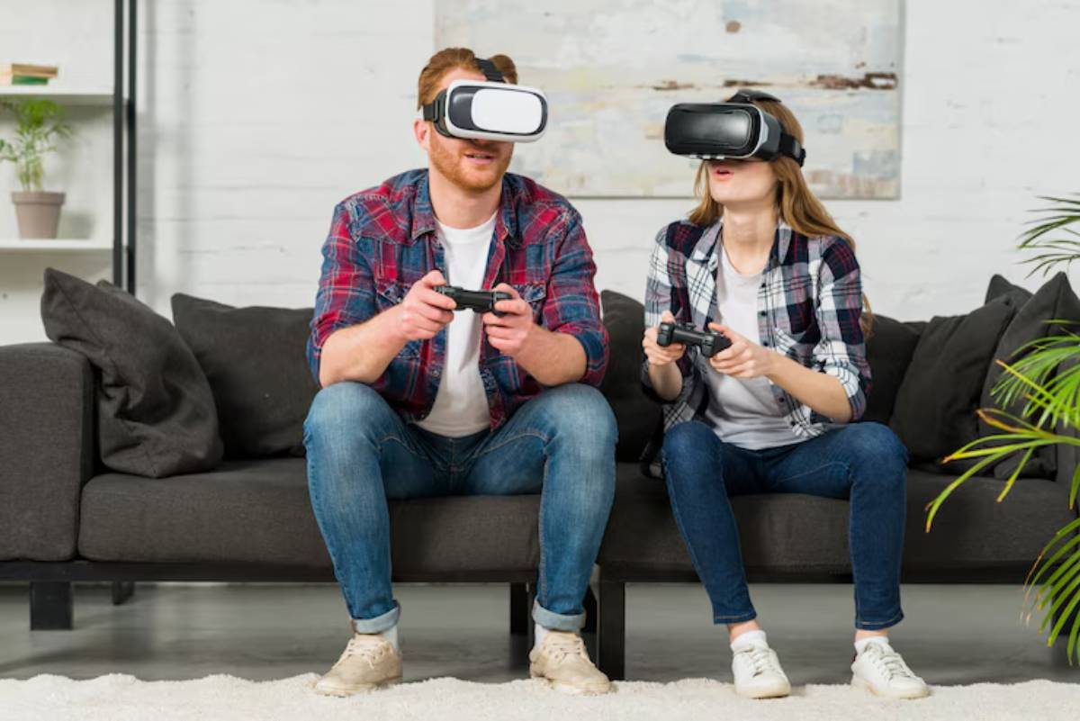 A man and a woman sit on a couch, each engaged in gaming; the man holds a controller while the woman wears a VR headset.
