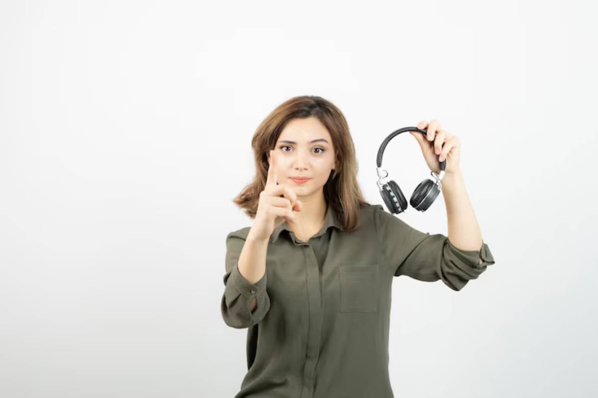 A person in a green shirt holds a pair of black headphones in one hand and gestures with the other, indicating attention.