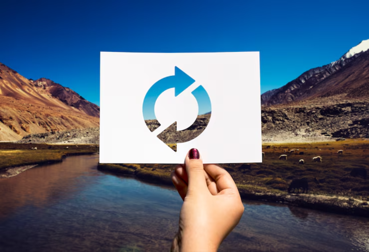 A hand holds a cut-out paper showcasing a recycling symbol against a scenic mountain landscape and serene river backdrop.
