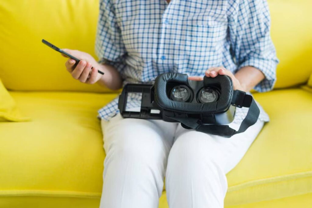 A person sits on a yellow couch holding a smartphone in one hand and a VR headset in the other, ready to explore virtual reality.