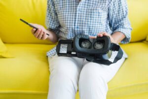 A person sits on a yellow couch holding a smartphone in one hand and a VR headset in the other, ready to explore virtual reality.