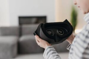 A child holds a black virtual reality headset, examining its lenses, with a cozy living room in the background.