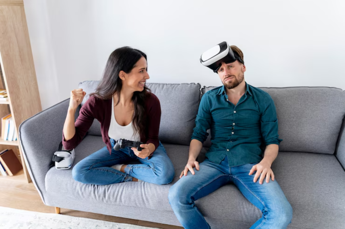 A woman sits cross-legged on a couch, excitedly holding a gaming controller, while a man beside her wears a VR headset.
