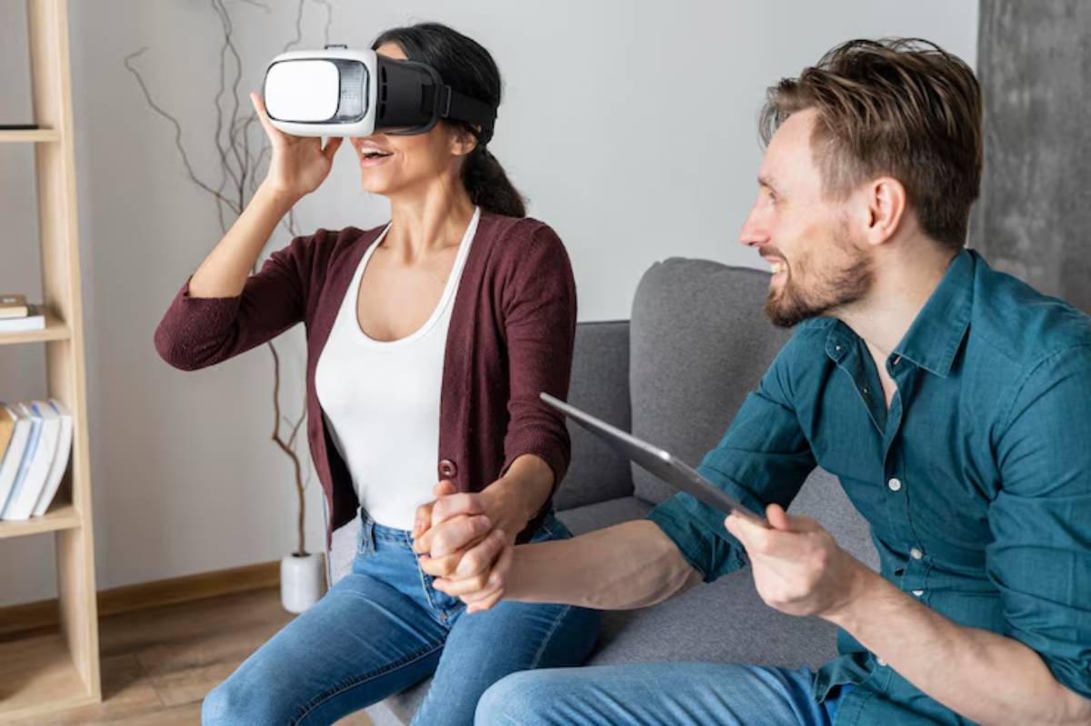 A woman wearing virtual reality goggles sits on a sofa, while a man beside her holds a tablet and supports her hand.