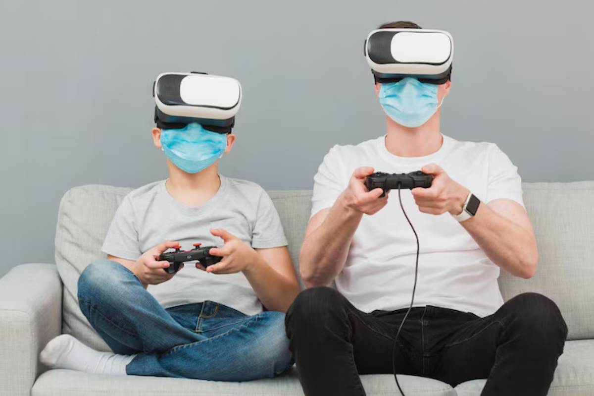 A child and an adult sit on a gray sofa, wearing virtual reality headsets and holding game controllers.