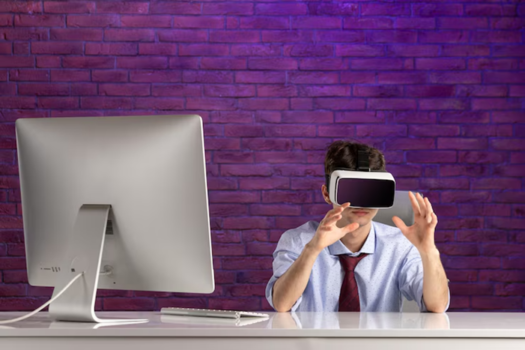 A person wearing a virtual reality headset is seated at a desk with a computer, surrounded by a purple brick wall backdrop.