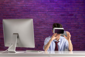 A person wearing a virtual reality headset is seated at a desk with a computer, surrounded by a purple brick wall backdrop.
