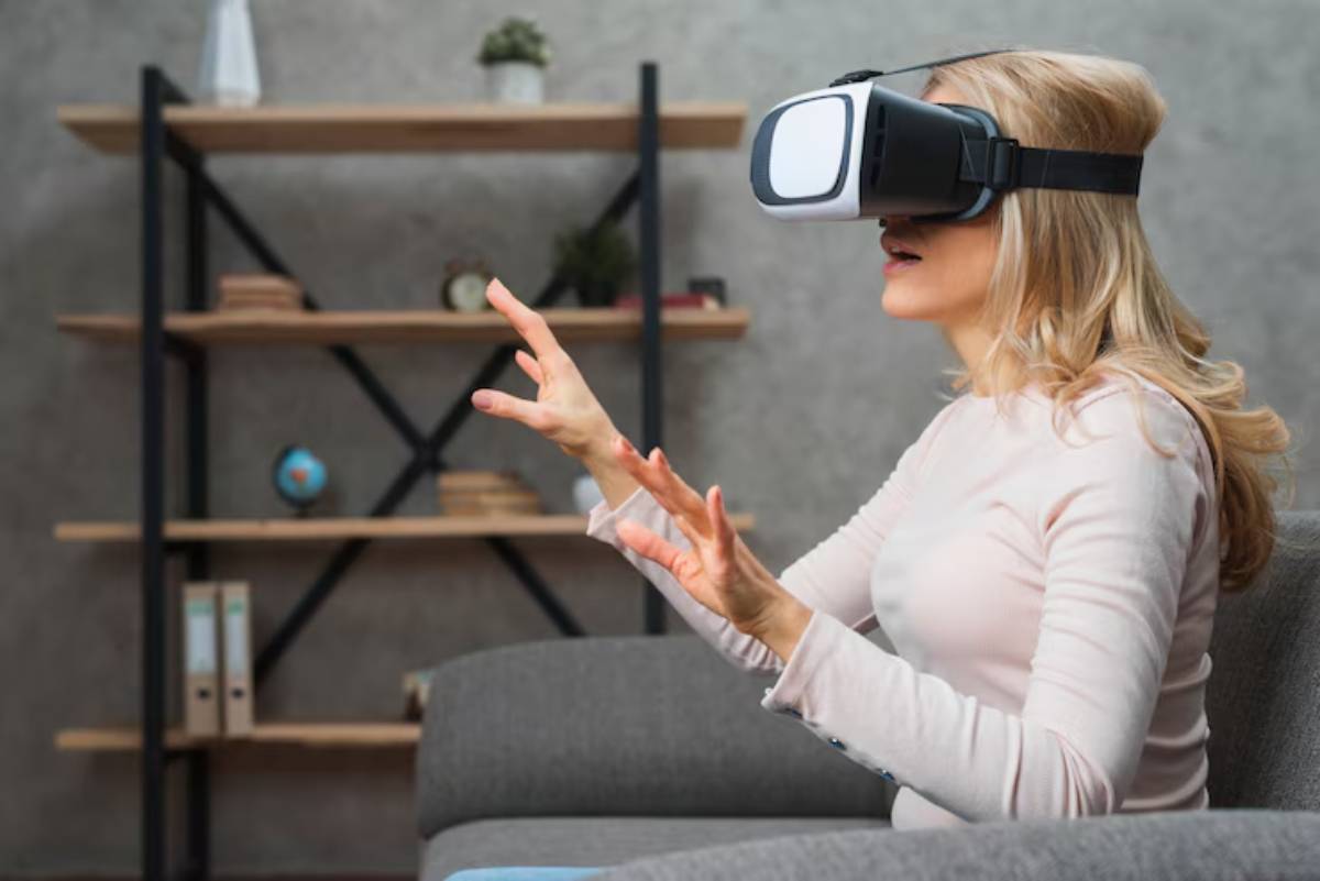A person sits on a gray sofa wearing a virtual reality headset, gesturing with both hands, with a modern shelf in the background.