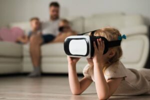 A young girl lies on the floor, immersed in a virtual reality headset, while family members relax on a couch in the background.