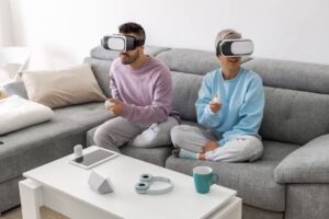 A couple sits on a gray couch, wearing VR headsets and holding controllers, surrounded by a tablet and headphones on a coffee table.