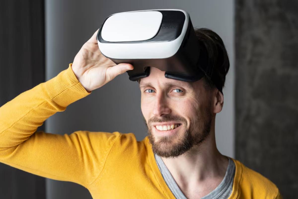 A person wearing a virtual reality headset, holding it up with one hand, dressed in a yellow sweater and gray shirt.