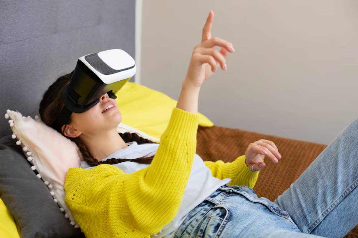 A person lying on a couch, wearing a VR headset, gesturing enthusiastically with one hand while relaxing on a yellow and brown blanket.