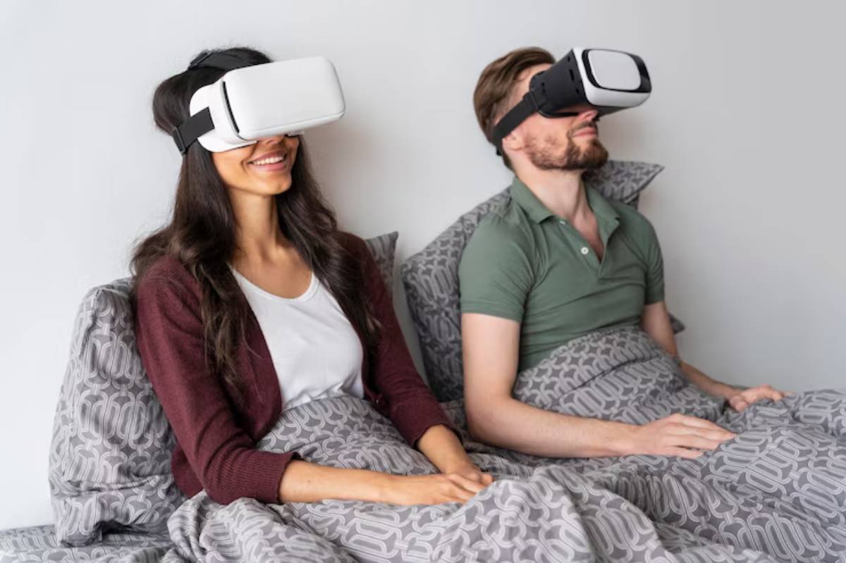 A man and woman sit side by side in bed, each wearing a VR headset, engaged in a virtual experience, covered in patterned blankets.