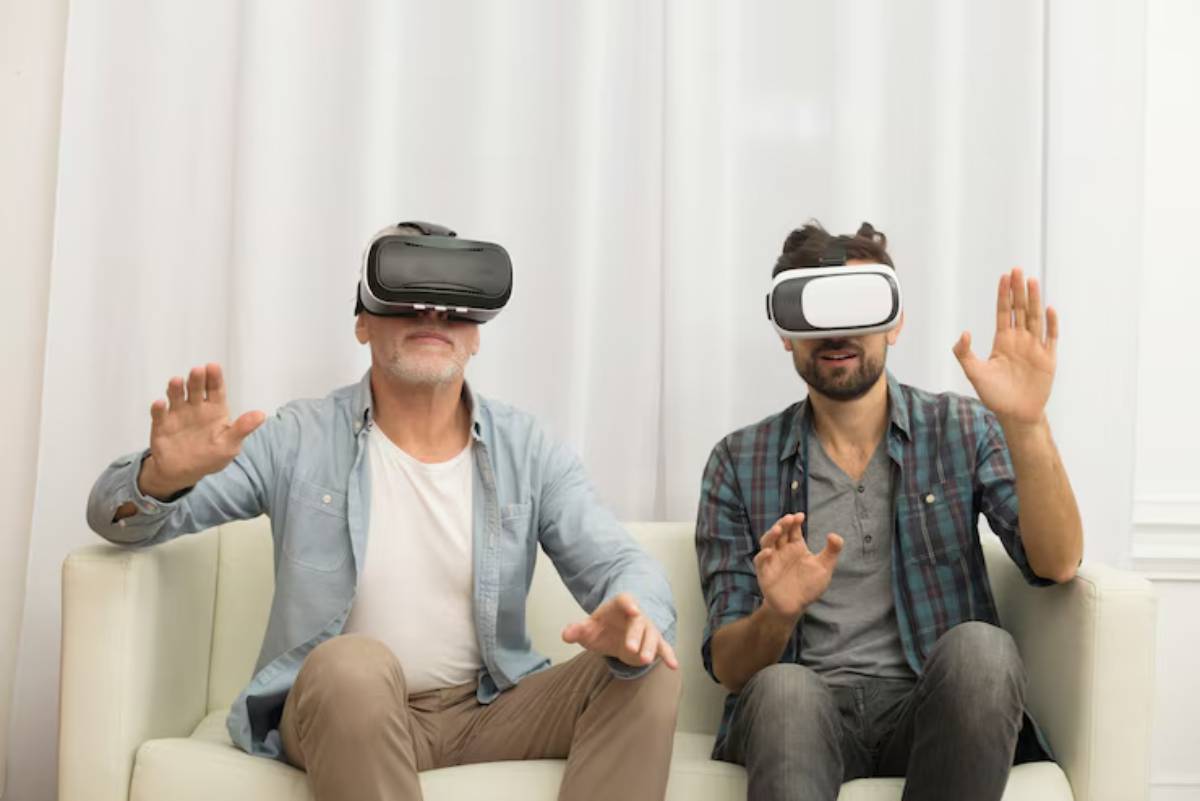 Two men wearing virtual reality headsets are seated on a couch, with one hand raised in excitement or interaction. White curtains in the background.