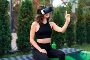 A woman in athletic wear sits on a bench, gesturing while wearing a virtual reality headset, surrounded by greenery.