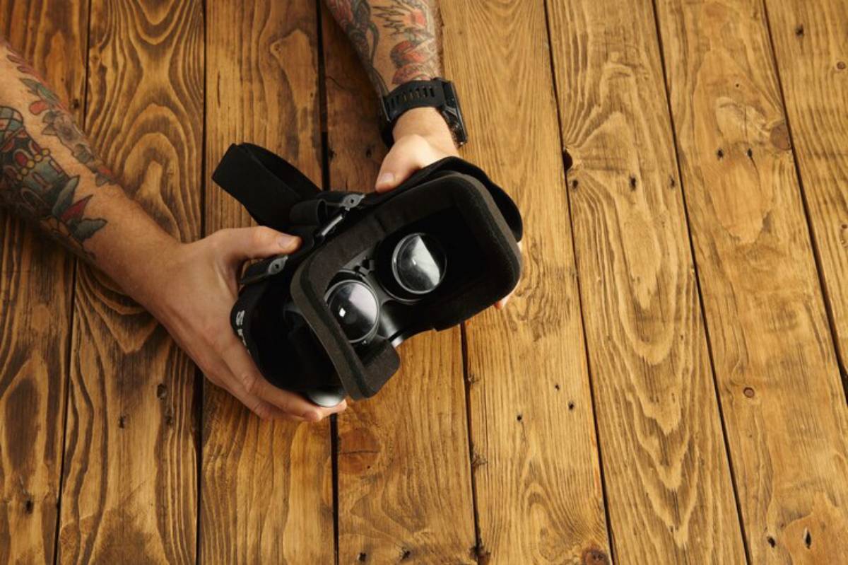A person with tattoos holds a VR headset over a wooden table, showcasing the device’s lenses and design.
