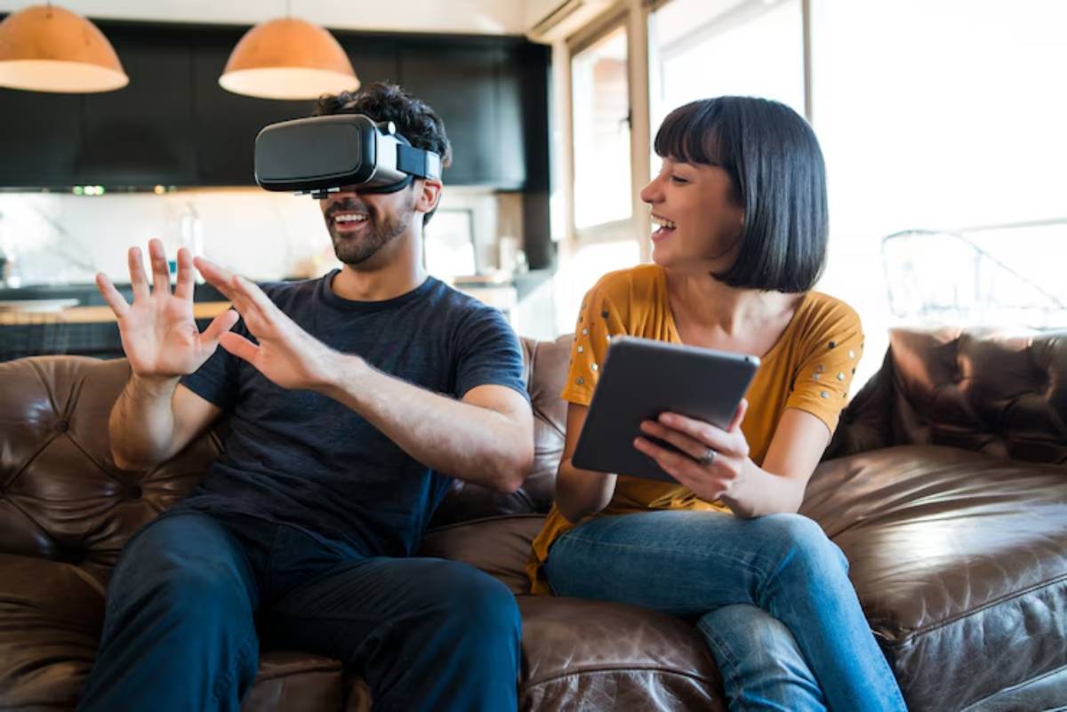 A man wearing virtual reality goggles gesturing animatedly while a woman sits beside him, holding a tablet on a couch.