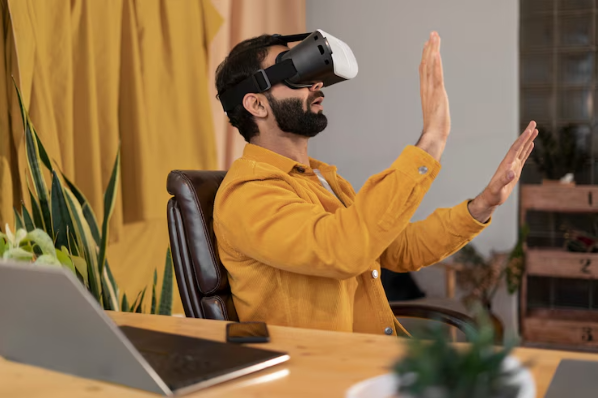 A person wearing a VR headset sits in a chair, arms raised in an interactive pose, surrounded by indoor plants and a laptop.