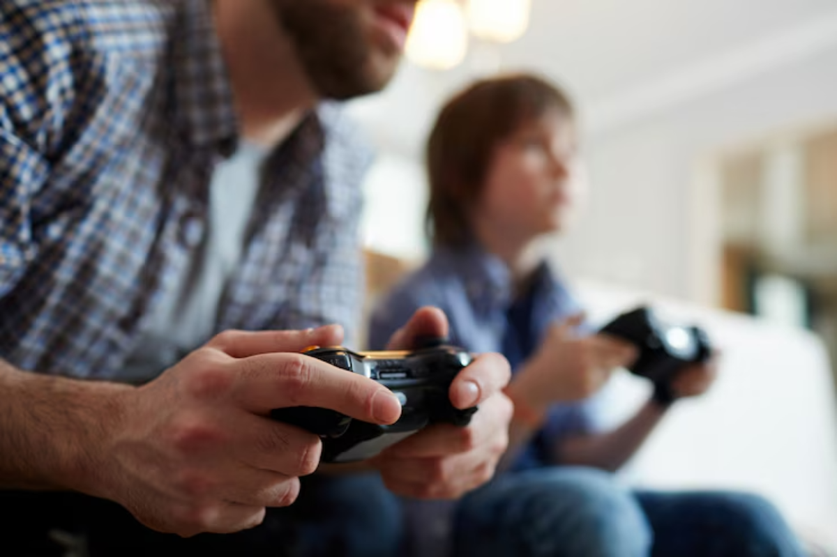 A man and child sit side by side on a couch, engrossed in video gaming, each holding a game controller.