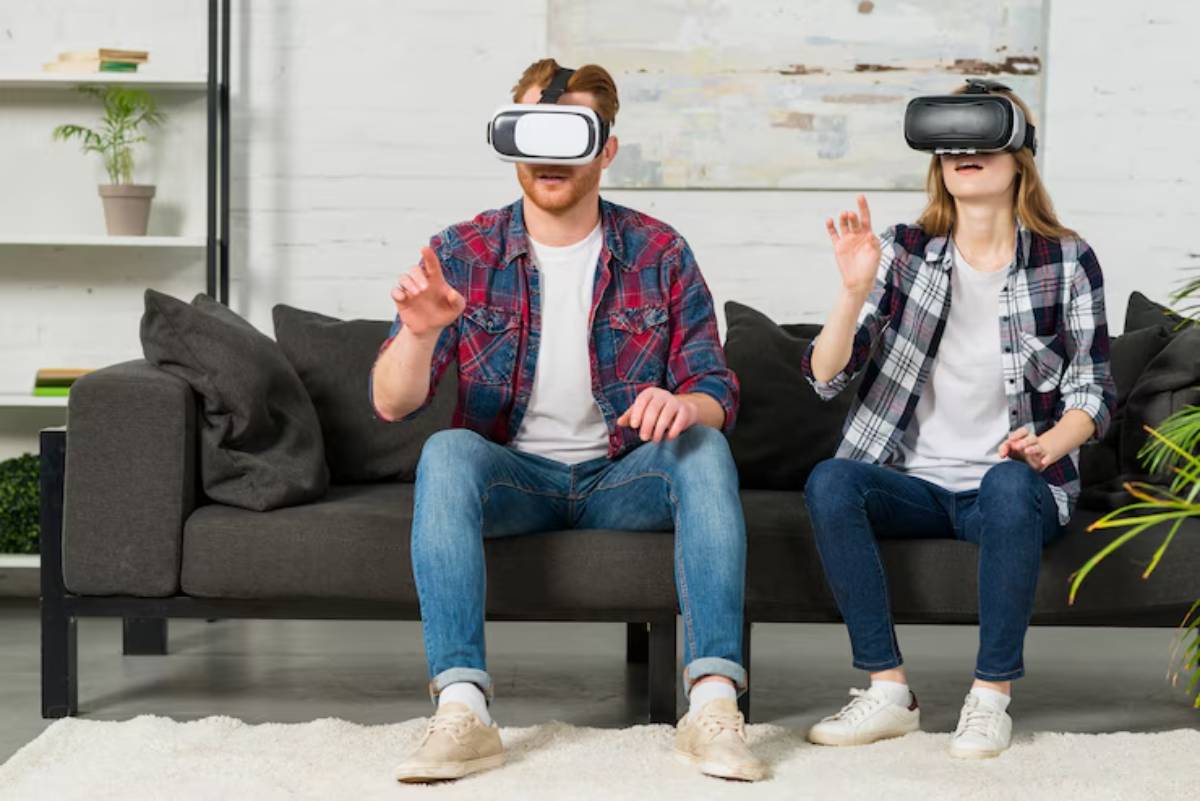 A man and woman wear virtual reality headsets while sitting on a couch, engaged in an immersive experience. Green plants in the background.