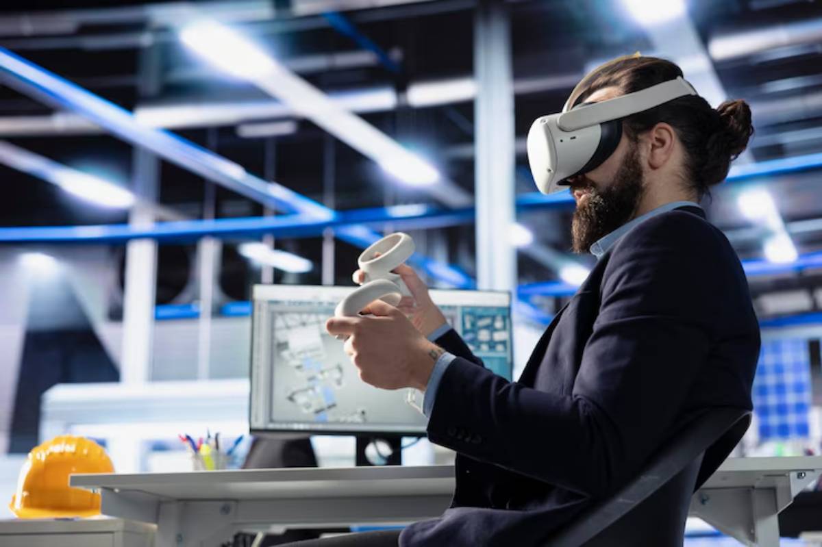 A person in a suit sits at a desk, wearing a VR headset and holding controllers, with a computer displaying technical plans in the background.