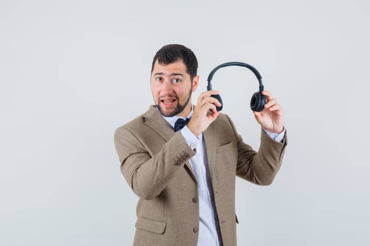 A man in a brown suit and bow tie holds a pair of black headphones, smiling enthusiastically against a light background.