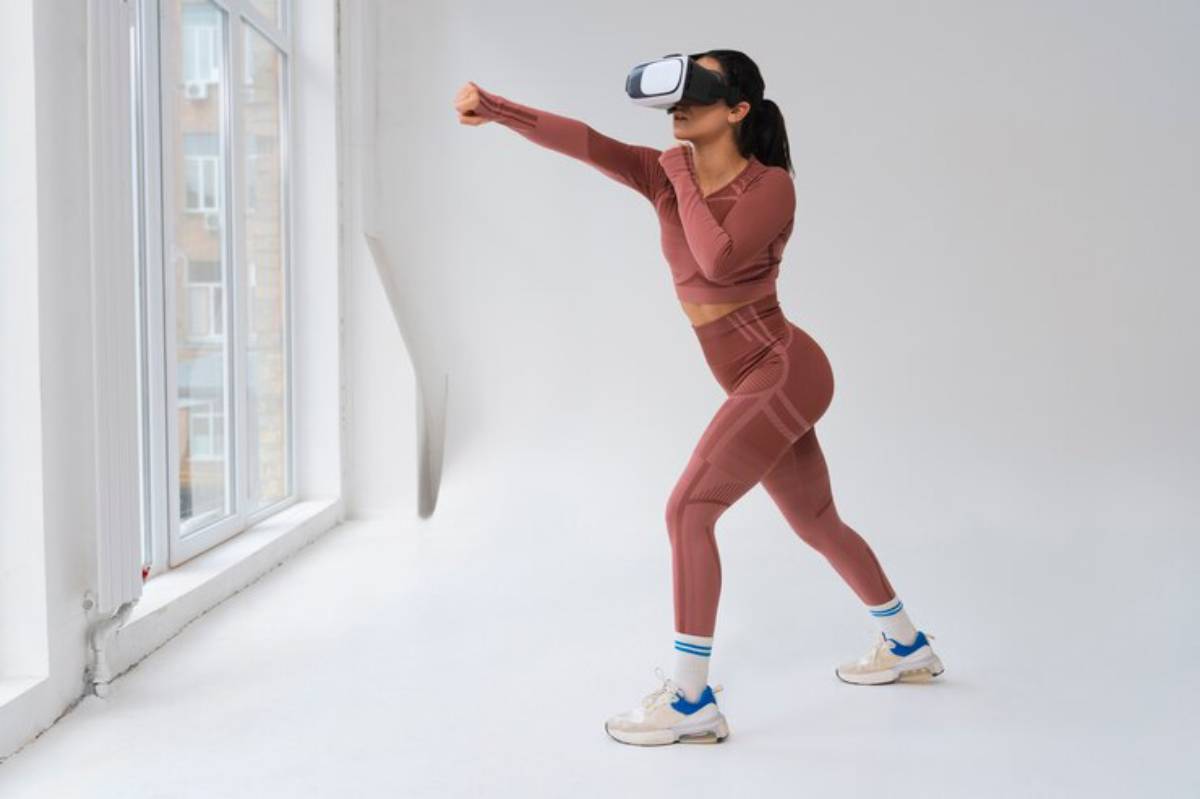 A woman in a pink workout outfit practices boxing moves while wearing a virtual reality headset indoors. Bright natural light floods the room.