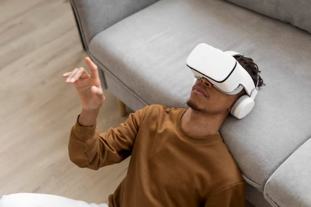 A person lies on the floor with a VR headset and headphones, gesturing while immersed in a virtual experience. A gray couch is nearby.