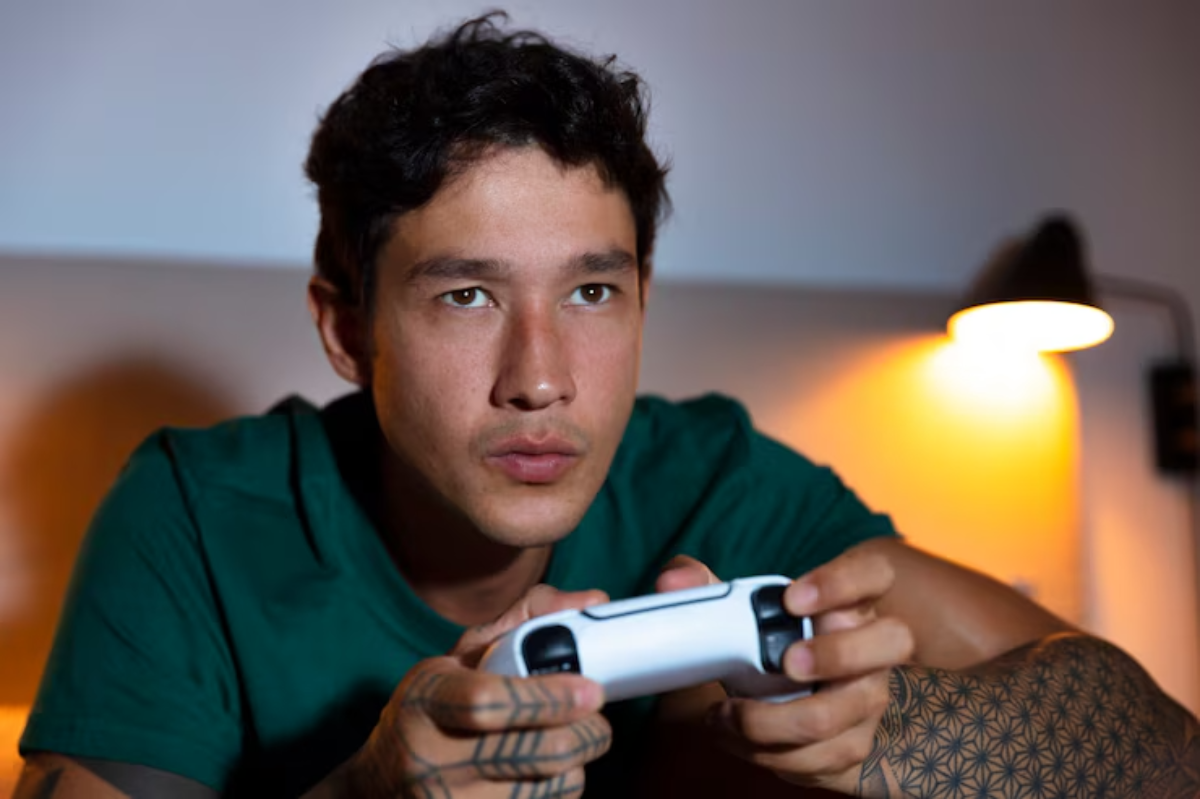 A person in a green t-shirt is focused on playing a video game with a white controller, illuminated by a warm lamp light.