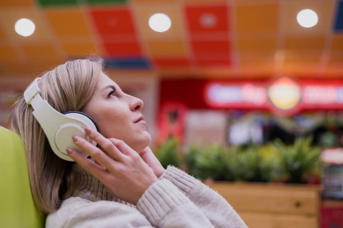 A young woman listens to music with white headphones, relaxing in a vibrant café filled with greenery.