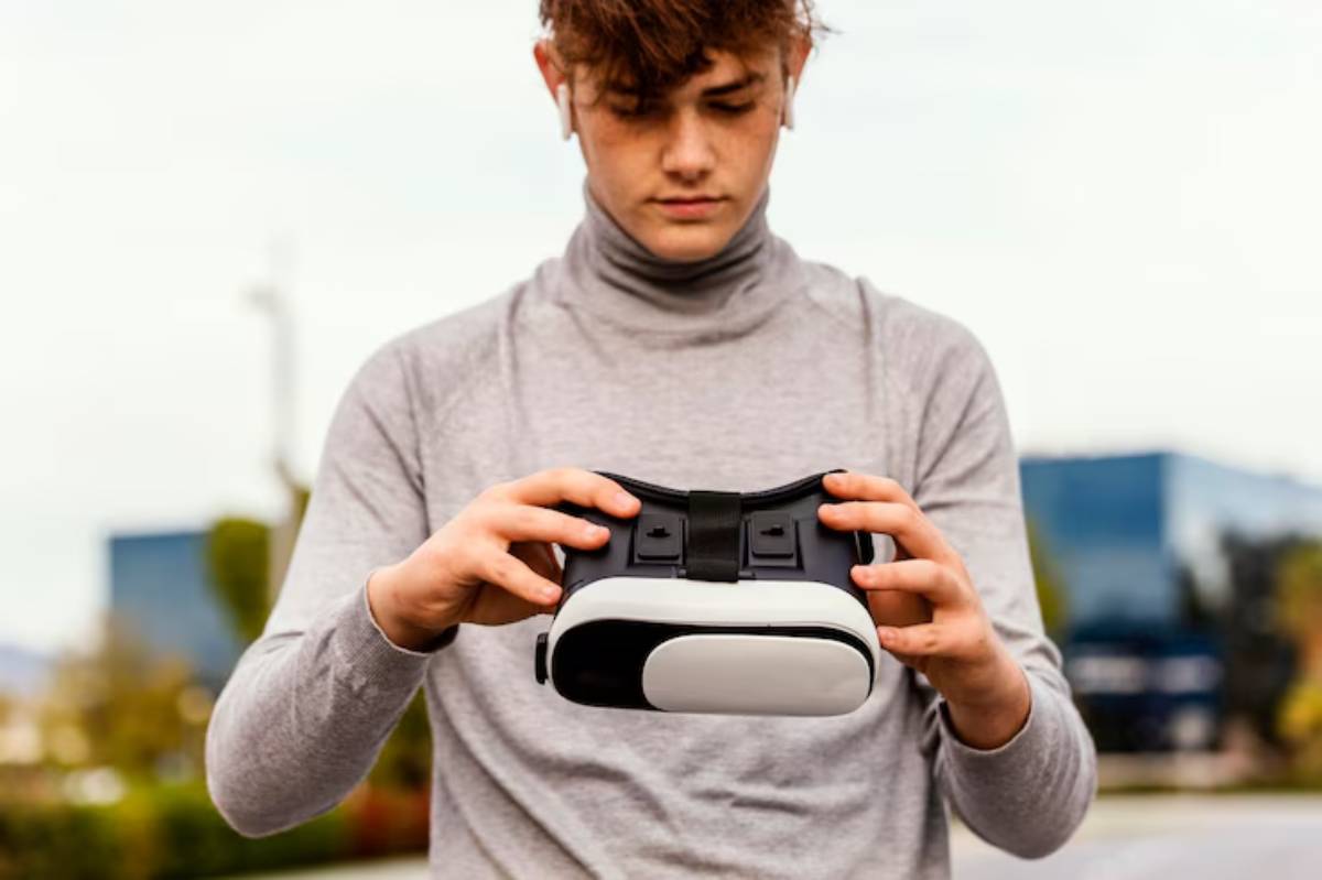 A person in a gray long-sleeve shirt holds a black and white virtual reality headset outdoors, with modern buildings in the background.