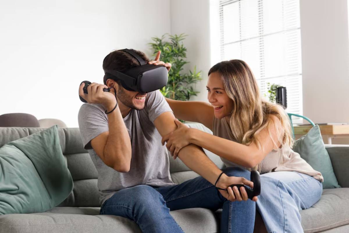 A person wearing a virtual reality headset sits on a couch while another individual playfully interacts with them.