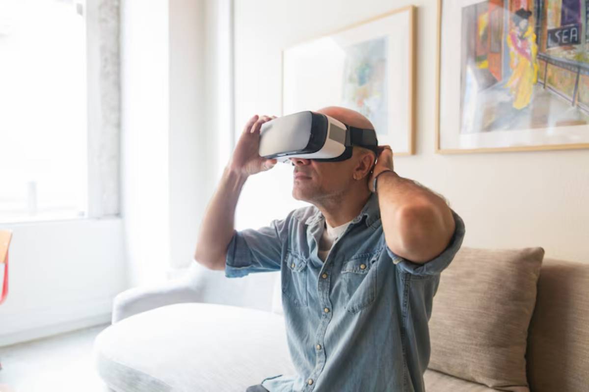 A man sits on a couch, adjusting a virtual reality headset, surrounded by bright natural light and colorful artwork on the walls.