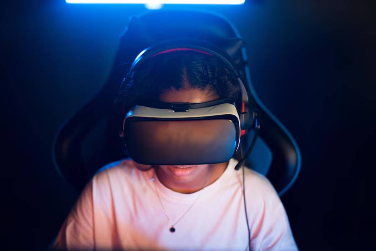 A gamer wearing a black headset sits in a gaming chair, illuminated by blue light in a dimly lit room.