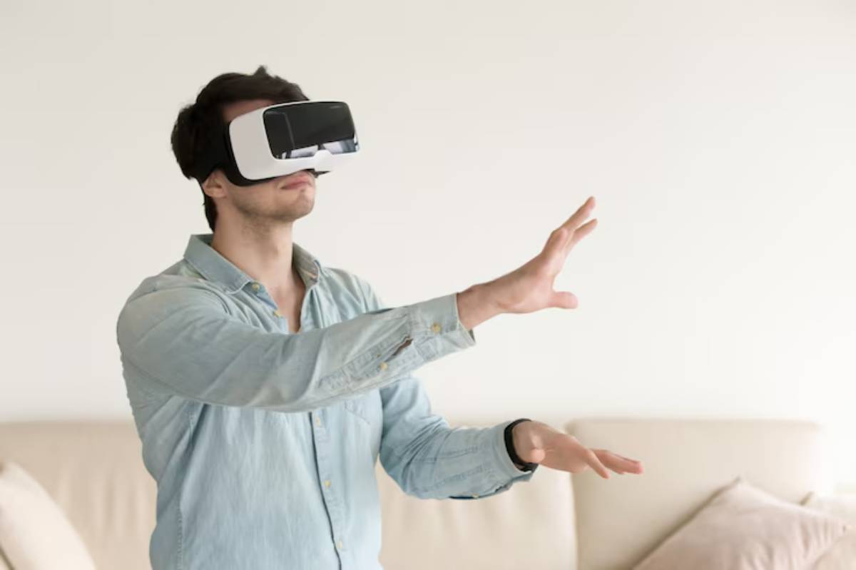 A person wearing virtual reality goggles is standing in a light room, reaching out with both hands as if interacting with a virtual environment.